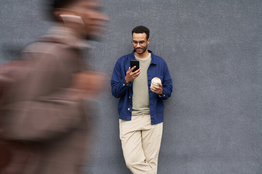 Young Black man standing against gray wall holding coffee cup and smartphone, looking at screen and smiling, blurred figure of another young Black man walking past in foreground