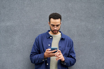 Young Black man standing against plain gray wall using smartphone, wearing glasses and casual clothing, looking down at device with focused expression, holding phone with both hands