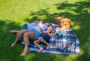 Mother hugs her sons while lying on a blanket during a summer picnic in a lush green park, enjoying a beautiful day outdoors, demonstrating closeness and quiet leisure time together