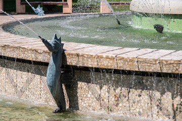 A copper fish sculpture in an urban water fountain with splashing water on a sunny day