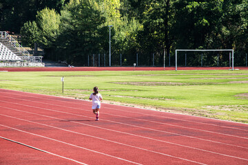 A little girl runs alone on the red tartan track of a small stadium during a sunny day