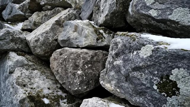 This close-up shot captures a rugged pile of large, weathered stones, showcasing intricate natural textures and a palette of grey and dark tones. Patches of white snow and delicate green moss and lich