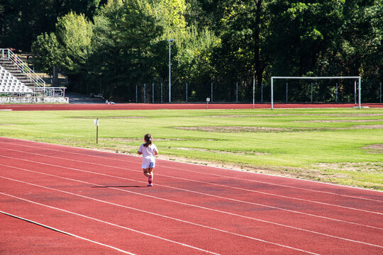 A little girl runs alone on the red tartan track of a small stadium during a sunny day