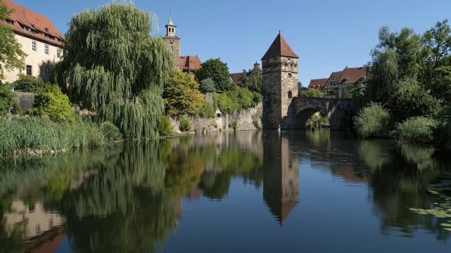 This picturesque scene captures the timeless beauty of an ancient riverside town under a clear blue sky. Historic stone buildings with traditional red-tiled roofs, including prominent towers and a cla