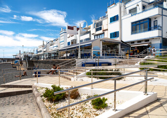 Puerto de las Nieves on Gran Canaria shows a sunny promenade with white houses, blue windows, seaside restaurants and a relaxed holiday atmosphere under a blue sky.