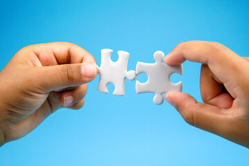 Two hands holding puzzle pieces facing each other on blue background. Concept of teamwork, partnership, connection, collaboration, unity, and problem solving in business or relationship.