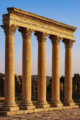 Naklejka premium Six ancient Roman Corinthian columns standing in a row at an archaeological site, captured in warm golden light with distant ruins and hills under a clear sky.