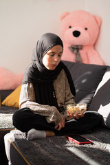Young girl in hijab sitting on sofa, holding a bowl of cereal and using a smartphone, cozy home setting with soft sunlight and teddy bear in background.