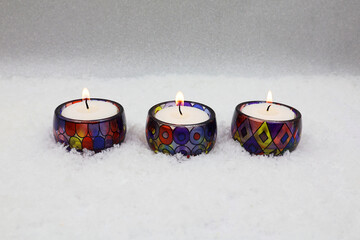 A row of burning candles in the snow. Tea lights in colored glass jars. A concept of atmosphere and coziness. Greeting card with copy space.