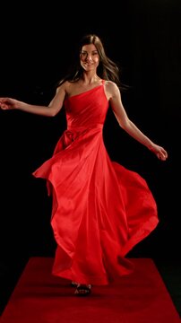 Stunning Black-Haired Woman in Ruby Red Dress and High Heels Gracefully Posing Against a Black Background, Captured in Super Slow Motion. Vertical Aspect Ratio. Speed Ramp Effect.