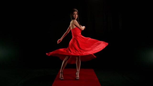 Stunning Black-Haired Woman in Ruby Red Dress and High Heels Gracefully Posing Against a Black Background, Captured in Super Slow Motion. Speed Ramp Effect.