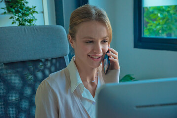 Professional woman engaged in phone conversation while working at desk with computer. office environment is modern and well lit, featuring plants and decorative items.