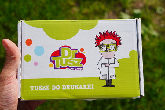 Hand holding a Dr. Tusz branded printer ink package featuring a cartoon scientist on a colorful background outdoors