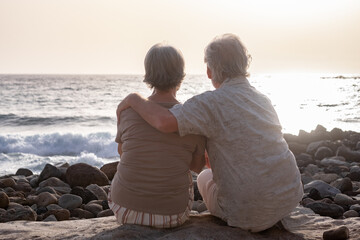 Fototapeta premium Back view of senior couple sitting on a rocky beach face the sea looking at the horizon and the sunset. Retirement lifestyle, vacation, freedom