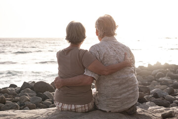 Fototapeta premium Back view of senior couple sitting on a rocky beach face the sea looking at the horizon and the sunset. Retirement lifestyle, vacation, freedom