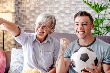 Cheerful couple of boy and elderly grandmother soccer fans watching football match on TV sitting on comfortable sofa in living room and expressing enthusiasm