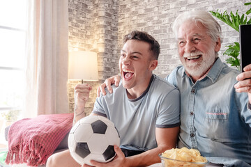 Excited young boy soccer fan watching a game on television together with his grandfather holding a soccer ball in hand sitting on a comfortable sofa in his living room