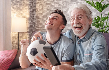 Excited young boy soccer fan watching a game on television together with his grandfather holding a soccer ball in hand sitting on a comfortable sofa in his living room
