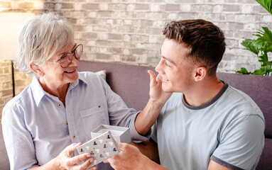 Young handsome boy gives his grandmother a birthday present while they are sitting at home on the sofa - taking care of the elderly. Grandparents day