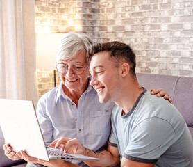 Elderly woman learning use of computer with her young nephew using together laptop for online connection sitting on sofa at home. Care and support towards the elderly we love