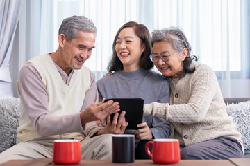 bonding happy asian family,mature adult daughter visit senior parents,sitting on sofa,relaxing,looking on digital tablet together in a cozy room