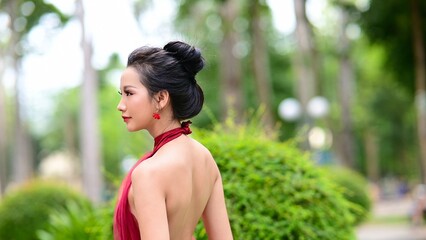Elegant Asian woman in red backless dress posing gracefully in green park under natural sunlight
