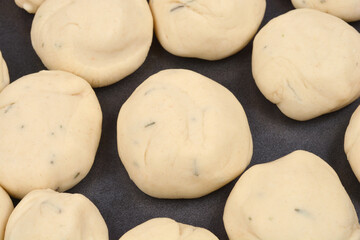 An overhead close-up shows numerous raw dough balls flecked with herbs arranged closely together on a dark baking sheet