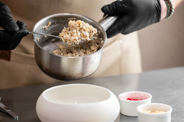 Chef scooping cooked grains from saucepan into bowl, with toppings nearby. Kitchen scene with apron, gloves, and stainless steel tools. Food preparation concept.