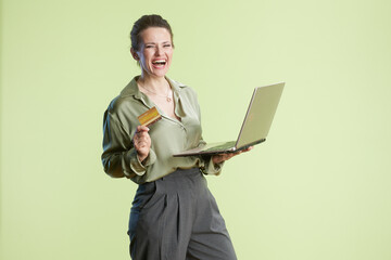 A laughing middle-aged woman holds a laptop and credit card, bathed in soft light. She is dressed in a green blouse and gray pants, enjoying the ease of online shopping.