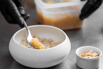 Close-up of gloved hands adding fruit compote to oatmeal in a white bowl. Healthy breakfast preparation with sliced almonds on stainless steel kitchen surface