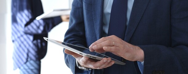 Businessman in a suit using a tablet during a corporate meeting, interacting with the touchscreen as blurred colleagues collaborate in the background, modern workplace tech