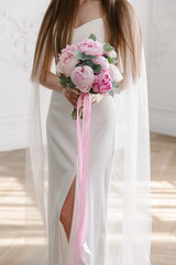 Elegant bride in a sleek white gown with a high slit holds a vibrant pink peony bouquet in a bright, sunlit minimalist studio interior