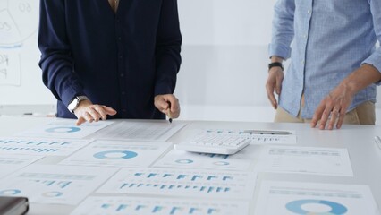 Business colleagues are examining financial data, pointing at charts and graphs spread out on a table during a collaborative analysis session in a modern office.