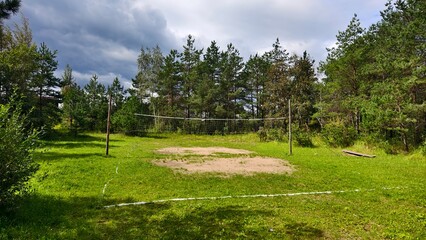 A sand volleyball court with wooden posts and a net between them sits on a grassy lawn surrounded by bushes, pine trees, and birch trees. It's a sunny summer day with a blue sky and clouds