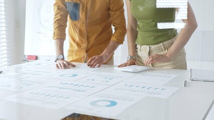 Business colleagues are working together, analyzing financial data and reports using a keyboard and printed charts spread out on a table in a modern office environment