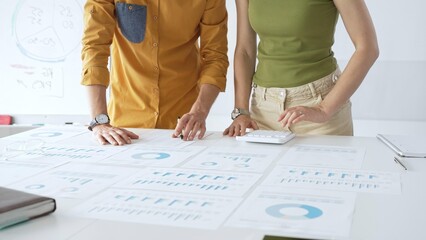 Professional team collaborating, reviewing financial documents and graphs on laptop with printed data spread across conference table in contemporary workspace. Business people concept