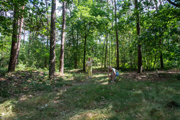 Two women enjoying a sunny summer hike in a lush green forest, one walking on a trail while the other crouches to explore foliage, relaxed and adventurous outdoors friends