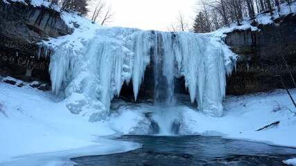 A frozen waterfall cascades amidst winter's grip, creating a frosty curtain over the water below. The scene is framed by snow-dusted cliffs and sparse trees - Powered by Adobe