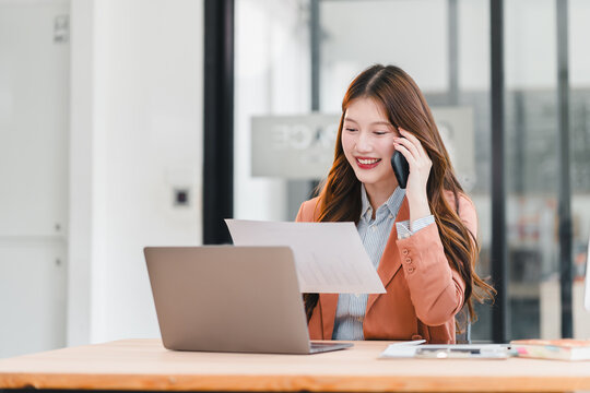 Young woman office worker smiling on phone while reading document at laptop in modern workspace