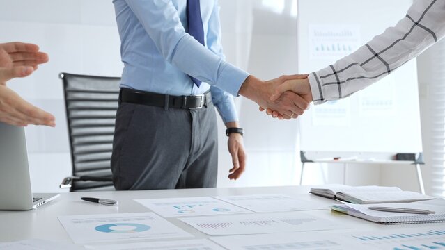Business partnership success concept with handshake in modern office, financial charts, graphs and laptop on desk representing teamwork, agreement and corporate growth