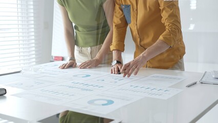 Two business professionals are examining financial data and reports, pointing at charts and graphs displayed on a table during a collaborative office meeting
