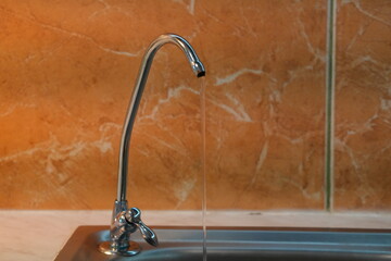 Modern Chrome Drinking Water Faucet with a Single Stream of Clear Water Flowing into a Stainless Steel Sink, Against an Orange Marble-Look Wall