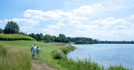Senior couple enjoying summer walk along a scenic lakeside trail, having leisure time outdoors, embracing togetherness and active retirement in nature under a blue sky
