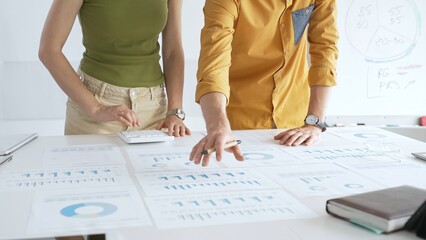 Professional team collaborating, reviewing financial documents and graphs on laptop with printed data spread across conference table in contemporary workspace. Business people concept