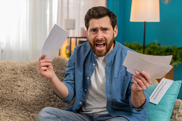Young angry man at home desperately hold a pile of unpaid bills on the sofa, feeling overwhelmed. Caucasian guy in frustration, struggling with financial debt and rising expenses.