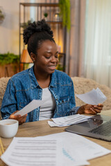 African American young woman sitting at table on home sofa nervously checks utility bills using laptop, printed papers. Black girl looks stressed overwhelmed trying to manage household payments online