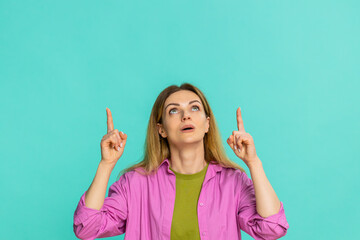 Middle-aged woman points finger upward with serious face, clearly signaling importance, asking to focus attention on something above. Girl isolated on blue background shows emphasis gesture and intent