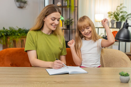 Mother and daughter at home draw grid on paper play tic tac toe fast rounds keep score in notebook. Woman and girl at table mark cross and noughts laugh plan rematch cheer friendly match track points.