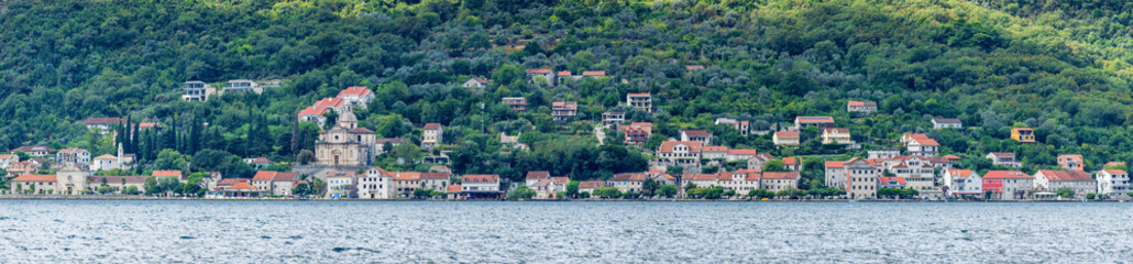Panoramic view of Prcanj, Kotor, Montenegro, along Jadranska magistrala with historic stone houses, church domes, and green hills rising above the calm Adriatic Sea.
