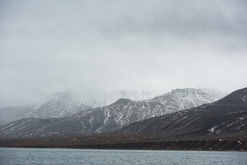 Fog drifts over snow-streaked mountains and a cold fjord shoreline on Svalbard, Norway, capturing the quiet beauty of the Arctic wilderness.
